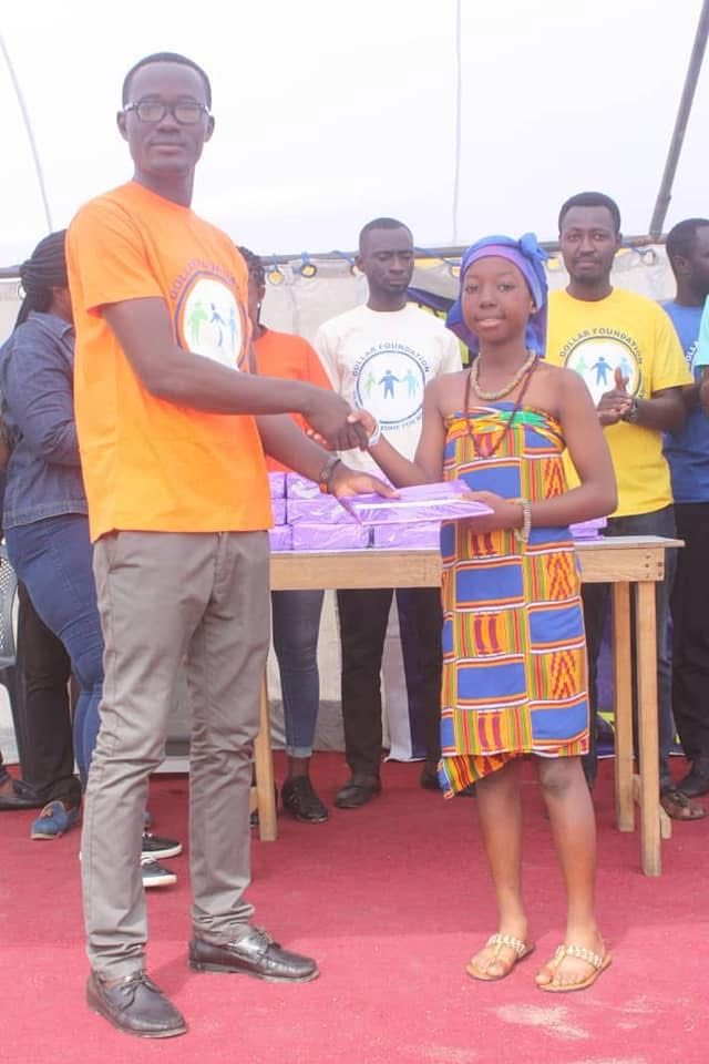 A volunteer shakes hands with a girl in kente cloth as he presents her with a wrapped gift.