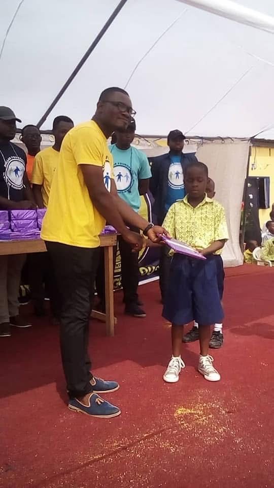 A volunteer in a yellow foundation T-shirt hands a wrapped gift to a young pupil on a red carpet.