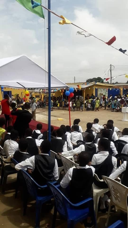 Pupils in white shirts and black waistcoats seated outdoors watching a school event, with bunting and balloons overhead.