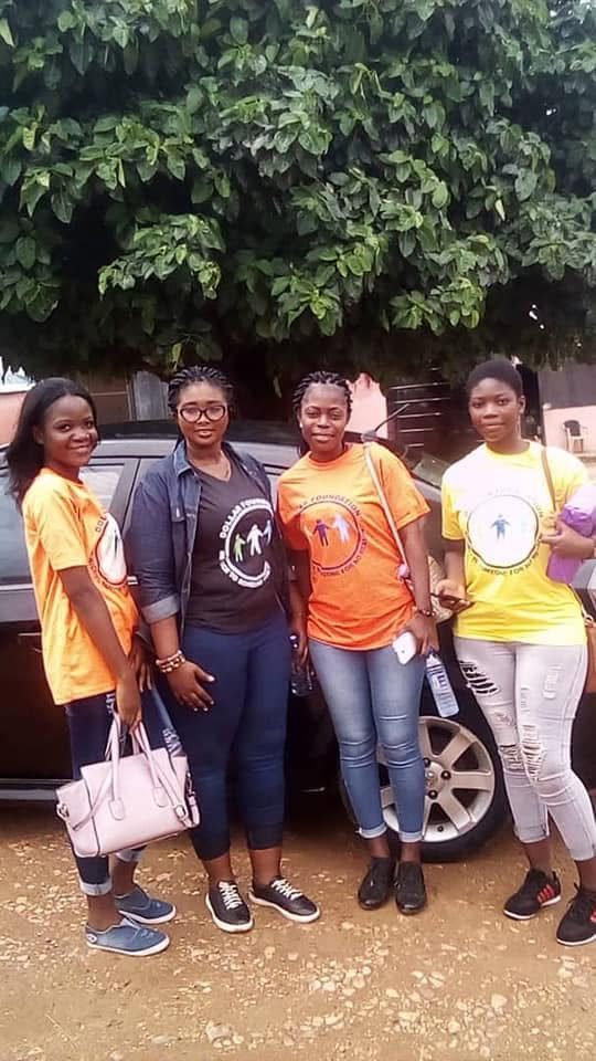 Four female volunteers in orange, grey and yellow foundation T-shirts standing in front of a car.