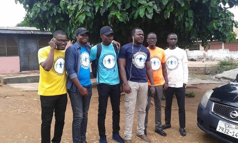Six male volunteers in yellow, blue, teal, navy, orange and white foundation T-shirts standing under a tree.