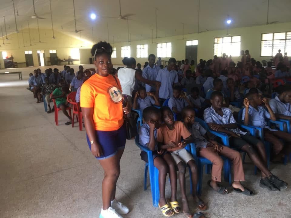 A volunteer in an orange foundation T-shirt stands beside seated students in the hall.