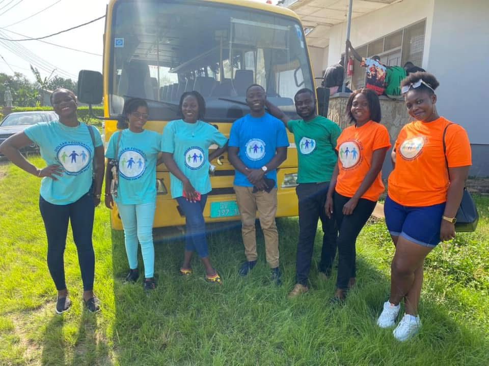 Seven volunteers in foundation T-shirts standing on grass in front of a yellow bus.