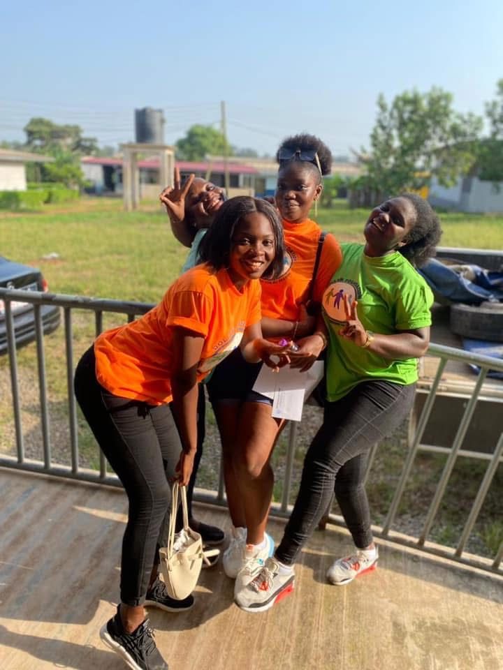Four volunteers in orange and green foundation T-shirts posing playfully on a veranda.