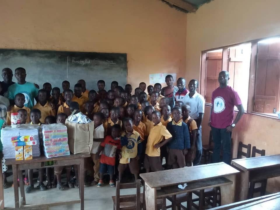 Pupils in yellow uniforms crowd around a table of exercise books and supplies, with volunteers at either side.