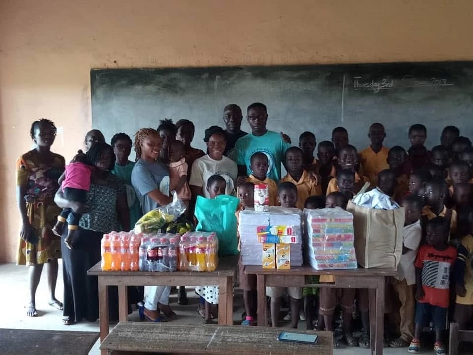 Volunteers, teachers and a large class of pupils behind tables of drinks, exercise books and other donated items in a classroom.