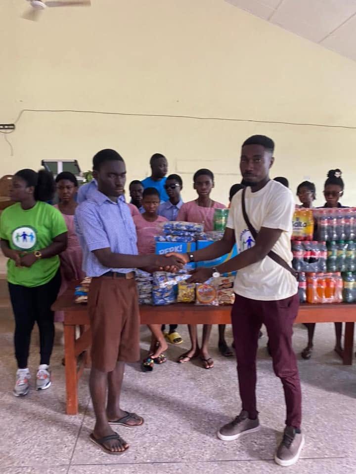 A Dollar Foundation volunteer shakes hands with a student in school uniform in front of a table stacked with drinks and packs of biscuits, as other students look on.