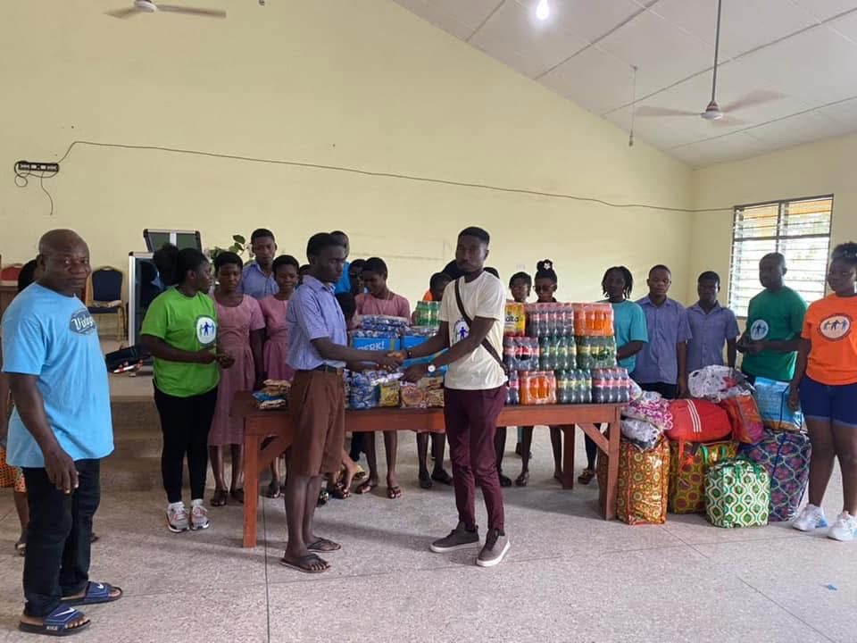 Students and volunteers in a hall around a table of drinks and biscuits, with bags of donated items on the floor.