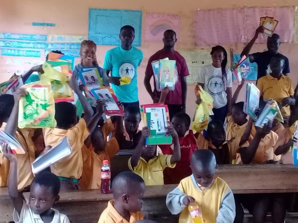 Pupils in school uniform hold up new exercise books and bags in a classroom, with volunteers standing behind them.