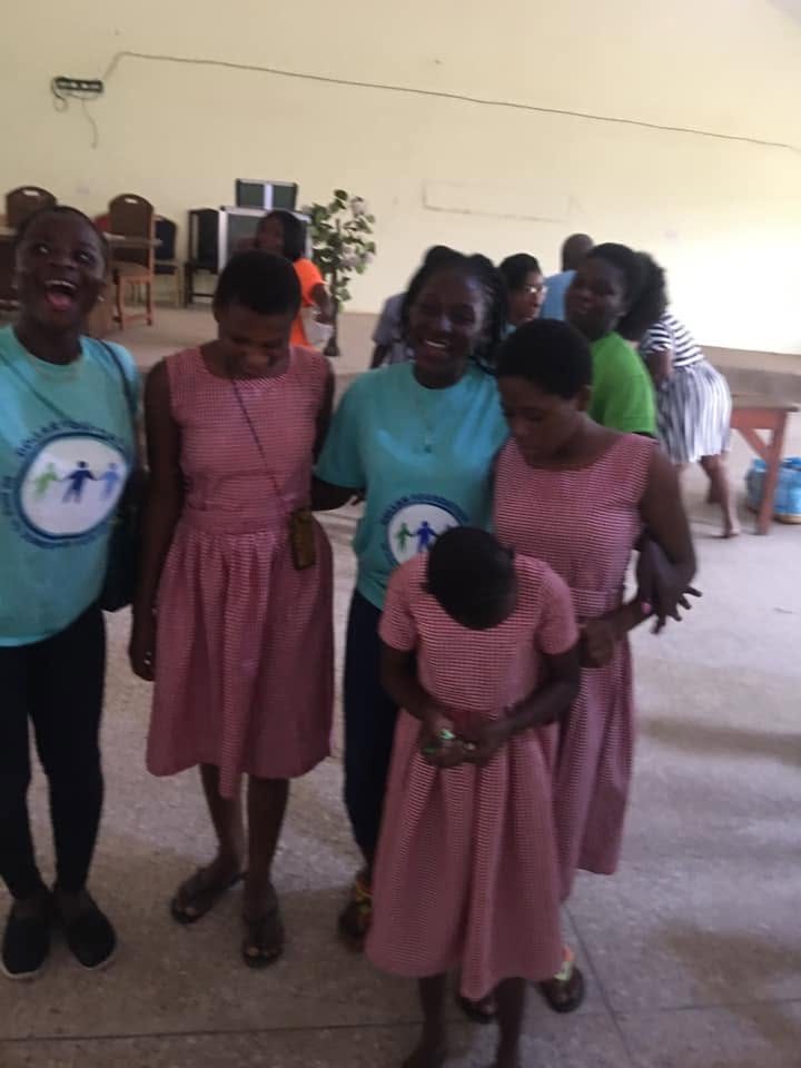 Two volunteers in teal foundation T-shirts laughing with schoolgirls in pink gingham uniforms.