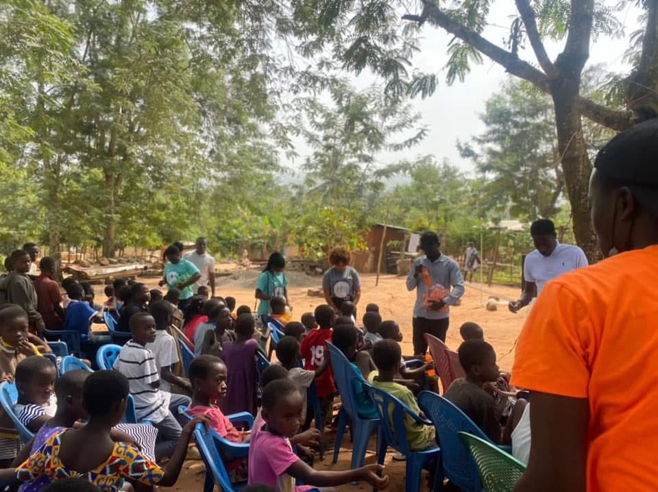 Children seated on plastic chairs under tall trees as volunteers stand and speak to them.