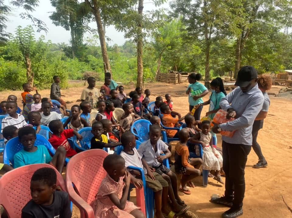 A volunteer hands out bottled drinks to children seated on plastic chairs outdoors.