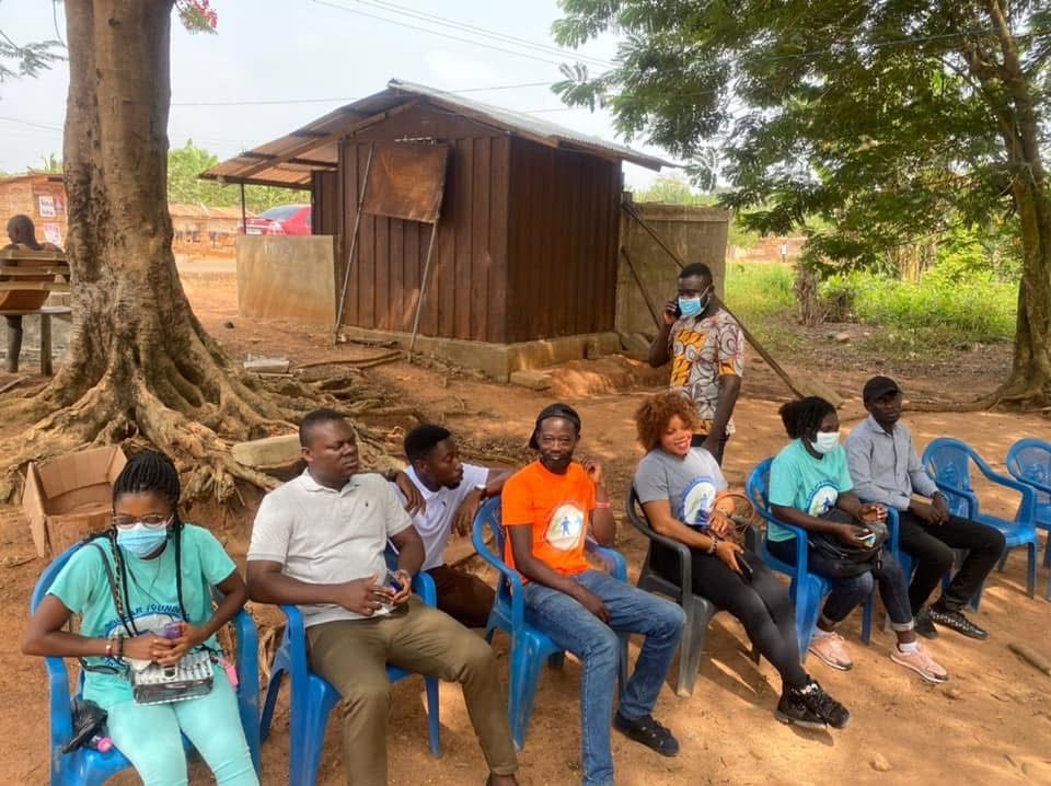 Volunteers, some wearing face masks, seated on blue chairs outdoors beside a large tree.