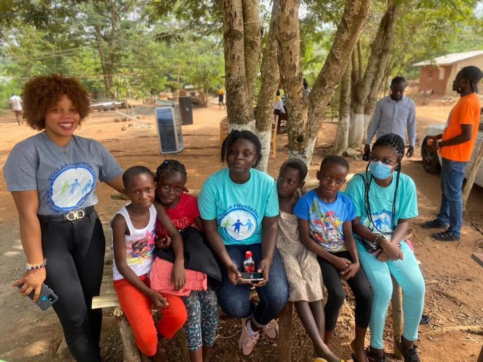 Two volunteers in foundation T-shirts sit on a bench under trees with five smiling children.