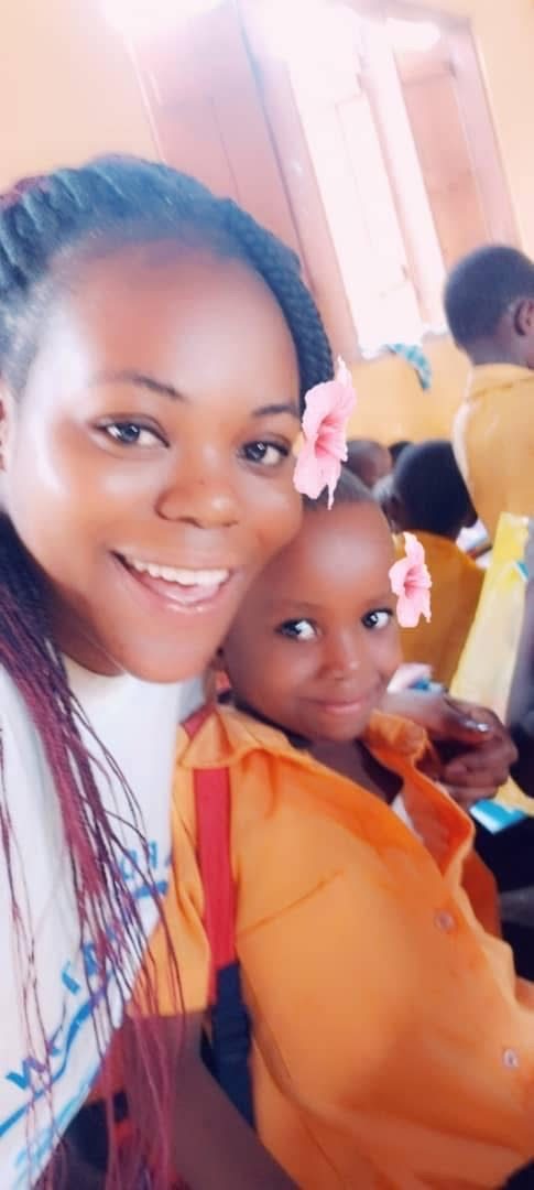 A smiling volunteer and a young pupil in an orange school uniform, both with pink flowers in their hair.